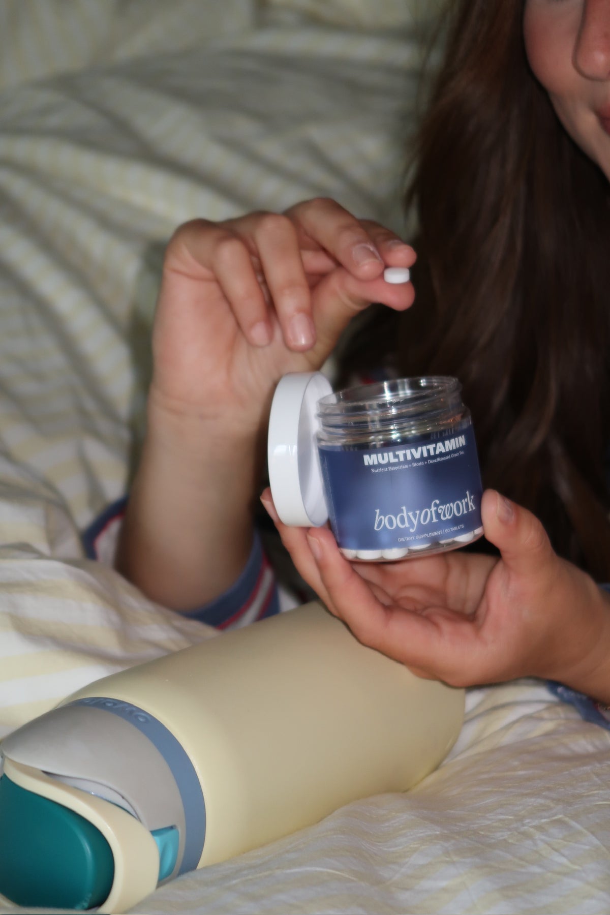 A person in bed holds an open jar of body of work Multivitamin tablets, with one hand dropping a white pill. A yellow water bottle is in the foreground on a white bedsheet background.
